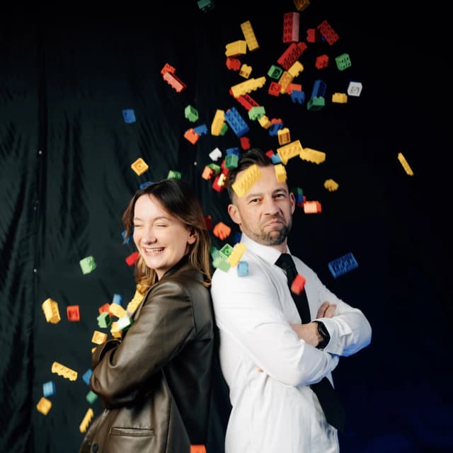 Zwei Personen stehen Rücken an Rücken vor schwarzem Hintergrund, während bunte Spielsteine durch die Luft fliegen – symbolisch für das kreative Duell beim Team-Duell Dresden