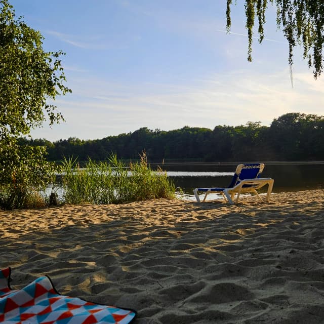 Liegestuhl auf einem Sandstrand am Brettmühlenteich in Thiendorf mit Blick auf den See und umliegenden Wald
