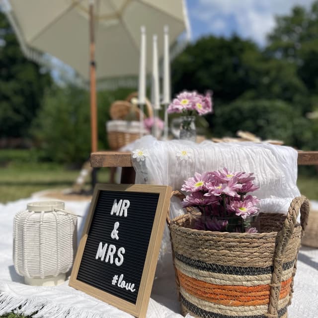 Detailaufnahme eines romantisch dekorierten Picknicks von Deluxe Picnic Dresden mit „Mr & Mrs“-Tafel, Blumen, Körben und Sonnenschirm im Hintergrund