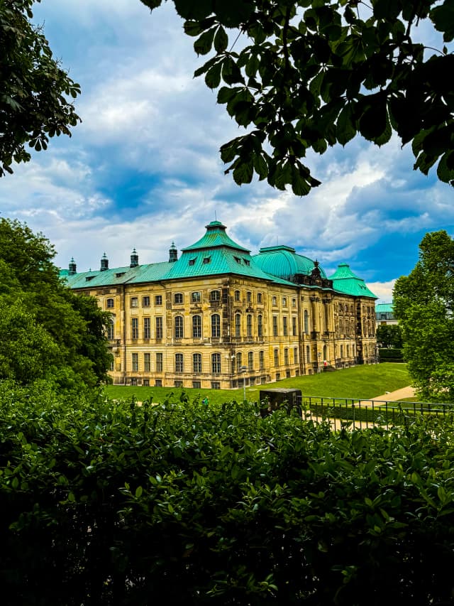 Seitenansicht des Japanischen Palais in Dresden, eingerahmt von dichtem Grün und Bäumen bei blauem Himmel